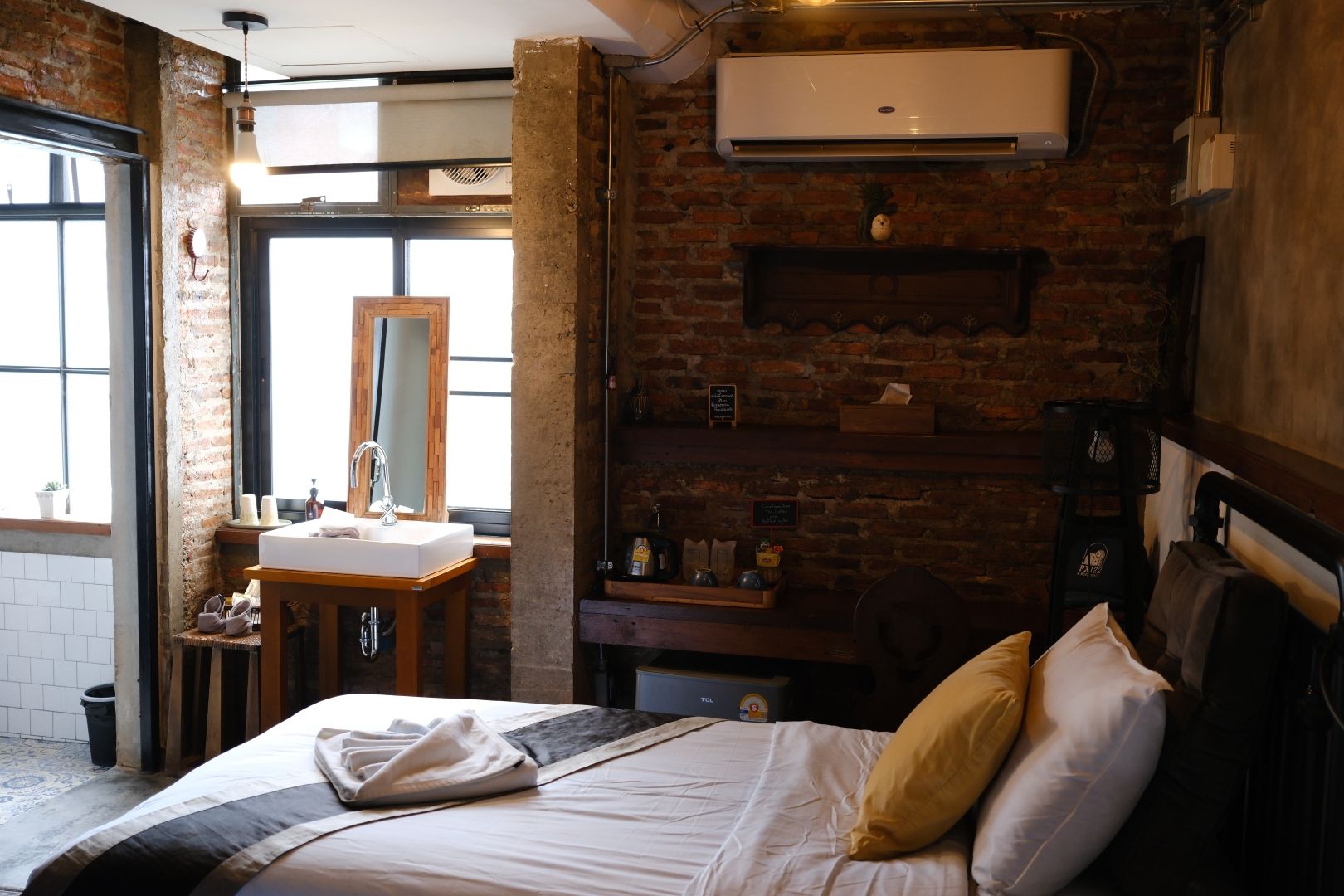 A hotel room with a brick wall, a large bed with pillows, a wooden nightstand with a mirror, a window, and a wall-mounted air conditioner.