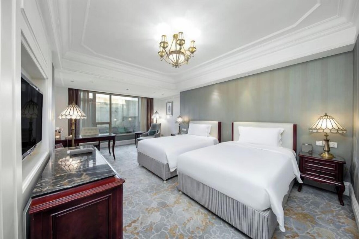 A hotel room with two beds, a desk, a TV, and a chandelier.