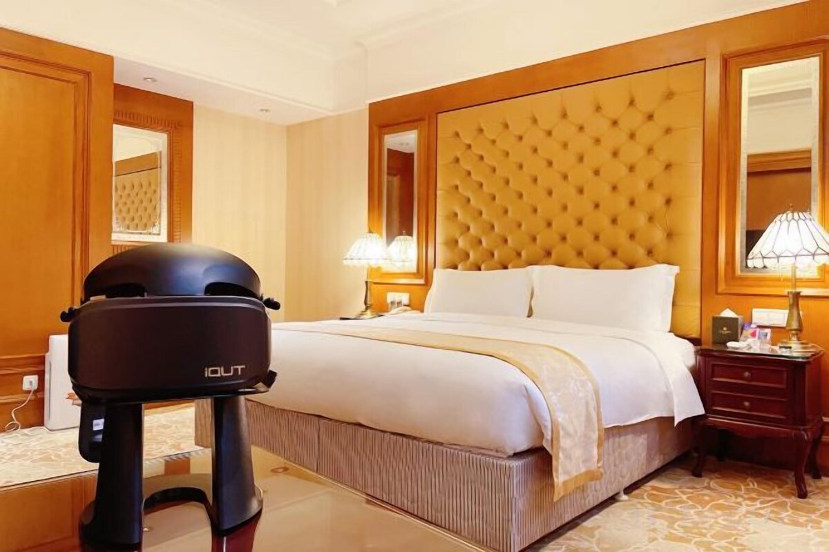 A hotel room with a large bed, a headboard, a nightstand with a lamp, and a small table with a device.