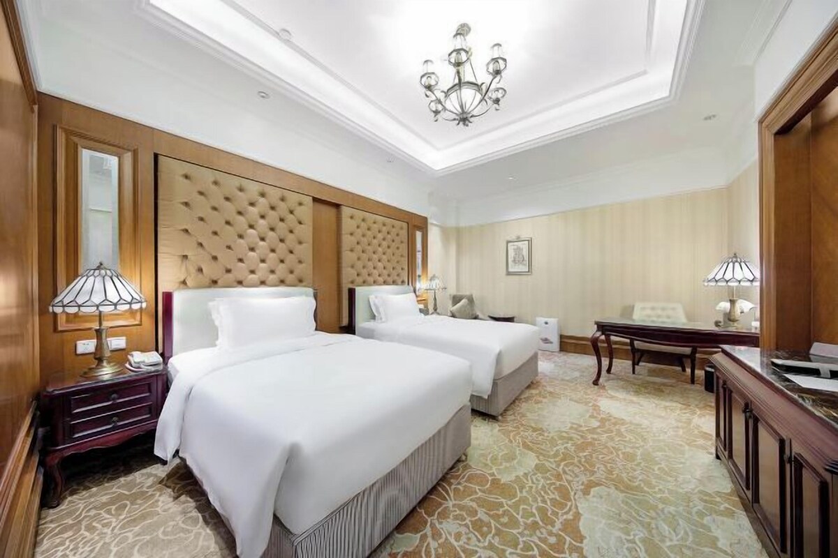 A hotel room with two beds, a chandelier, a wooden headboard, a desk, and a chair.