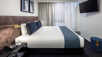 Une chambre d’hôtel avec un grand lit, une table de chevet avec un téléphone, un téléviseur à écran plat fixé au mur et une fenêtre avec des rideaux.