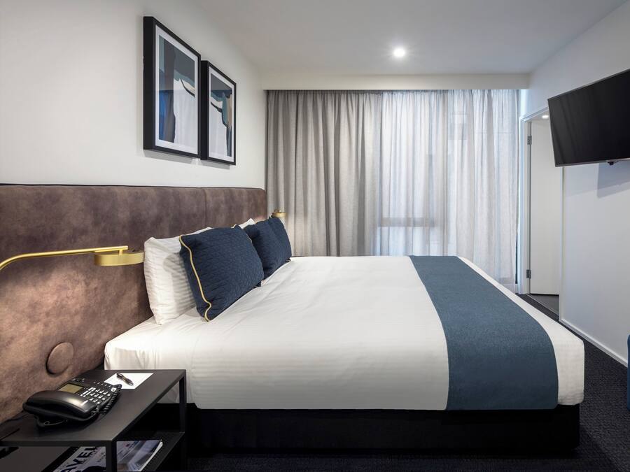 Une chambre d’hôtel avec un grand lit, une table de chevet avec un téléphone, un téléviseur à écran plat fixé au mur et une fenêtre avec des rideaux.