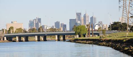 A city skyline with modern buildings, a bridge over a river, and a park area with trees and a grassy field.