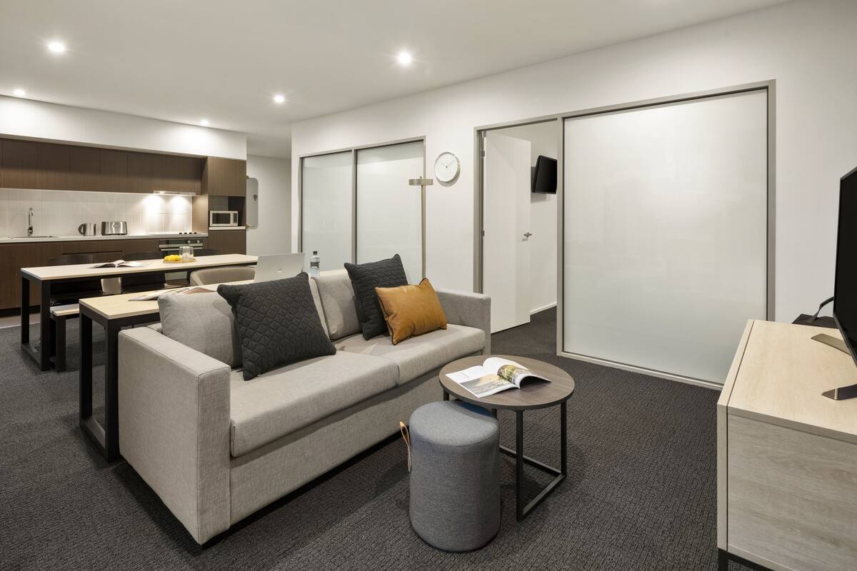 A modern living room with a sofa, coffee table, and a small ottoman. There is a kitchen area with a microwave and a dining table. The room has a neutral color scheme and a large sliding door closet.