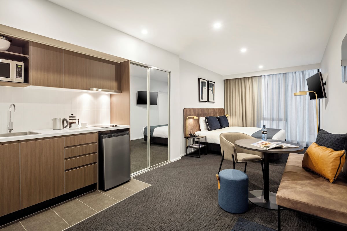 A modern hotel room with a kitchenette, a bed, a seating area, and a TV.