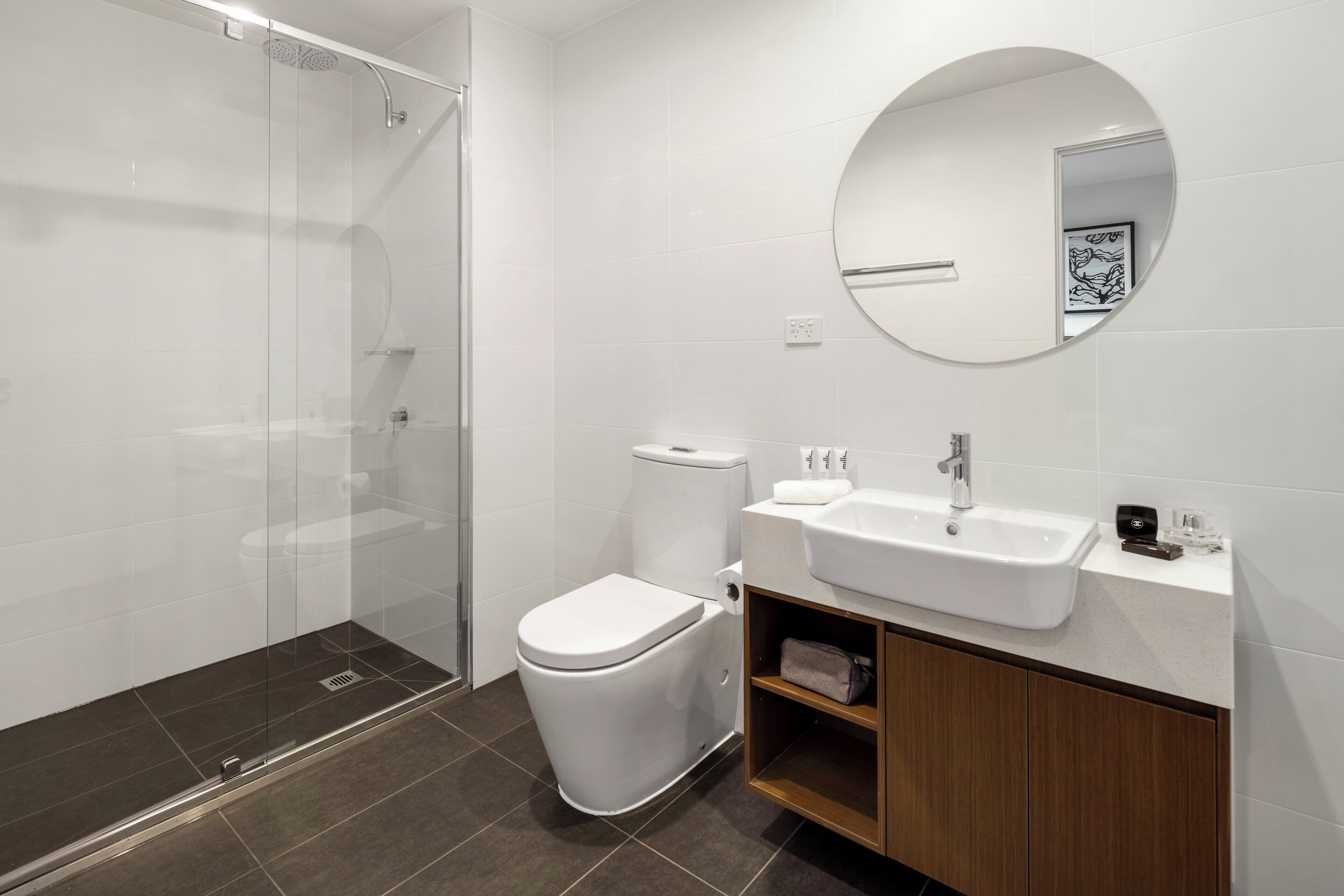 Un baño moderno con ducha de vidrio, inodoro blanco, lavabo con espejo y mueble de madera.