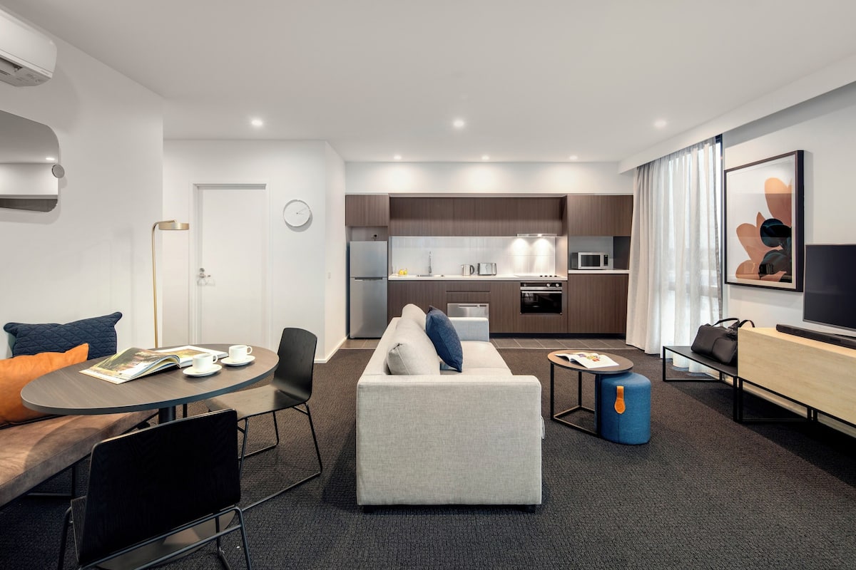 A modern hotel room with a living area, a dining table, a sofa, a small kitchen, and a TV.