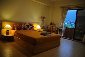 Een slaapkamer met een groot bed, twee bedlampjes, een kleine tafel met een lamp, een stoel en uitzicht op een bergachtig landschap door een schuifdeur.