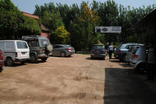 A parking area with several vehicles, including a white van and a silver SUV. There is a sign for City Gaon Restaurant in the background. - City Gaon Resort (Suryabinayak)