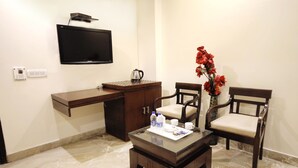 A hotel room with a desk, two chairs, a coffee table, a TV mounted on the wall, and a vase with red flowers. - Hotel Paradise Inn (New Delhi)