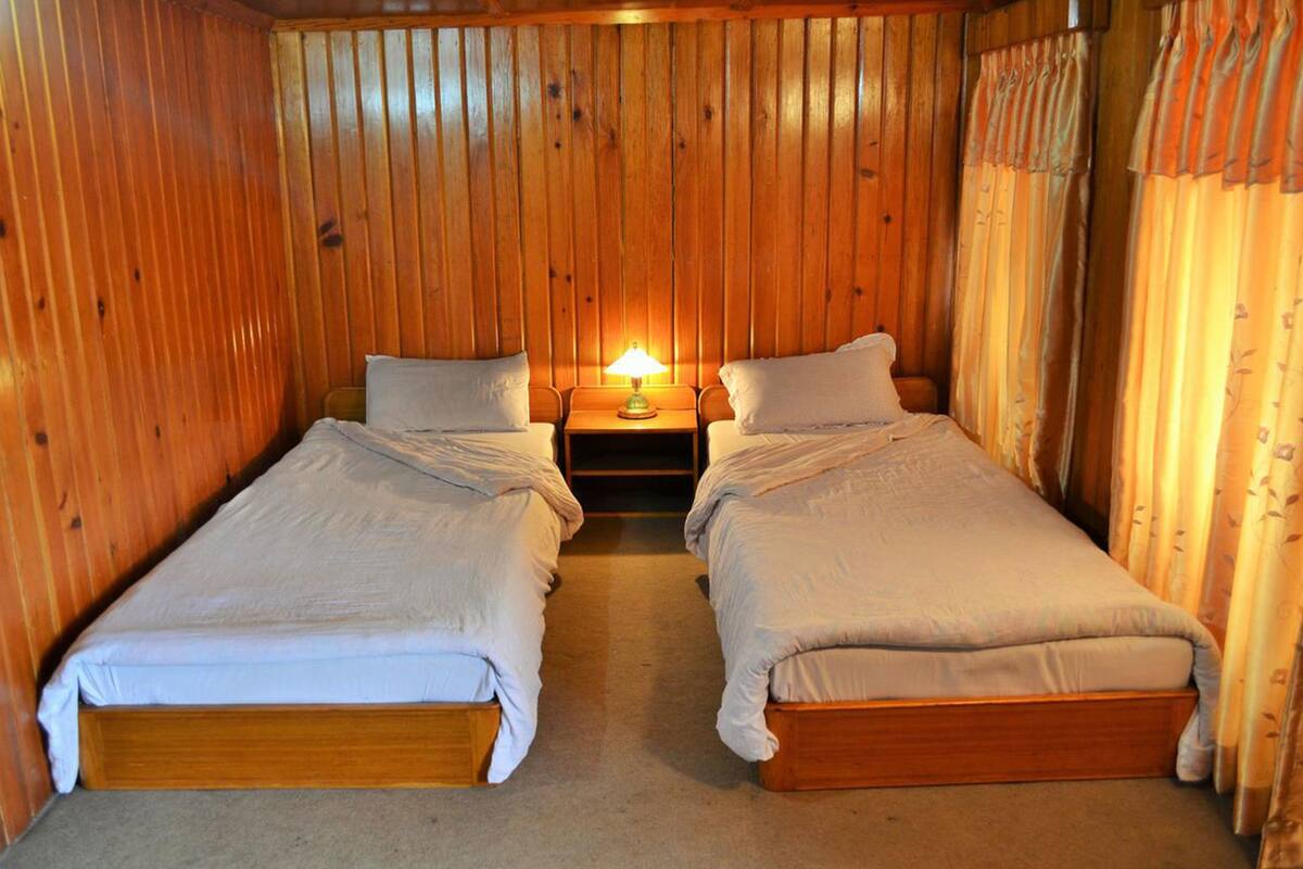 A room with two single beds, a nightstand with a lamp, wooden walls, and curtains.