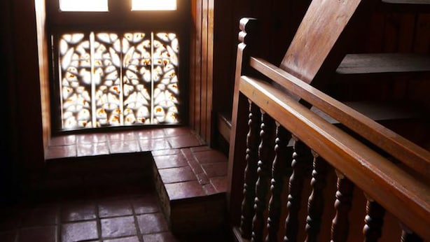 A wooden staircase with a handrail, leading to a room with a window featuring a stained glass design. - Unique Guest House (Bhaktapur)