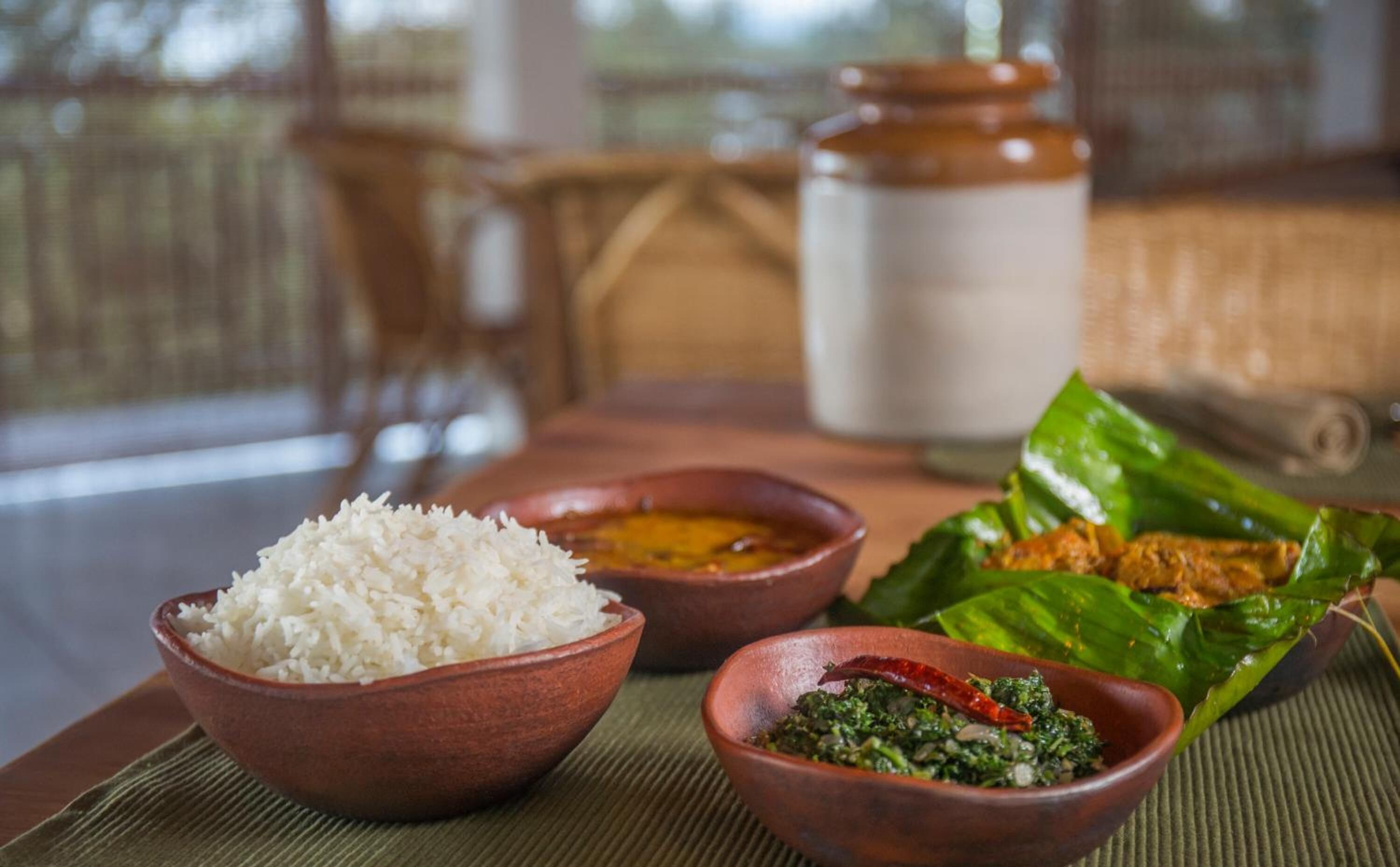 Une table avec un bol de riz blanc, un bol de légumes verts et une assiette de nourriture posée sur une feuille.