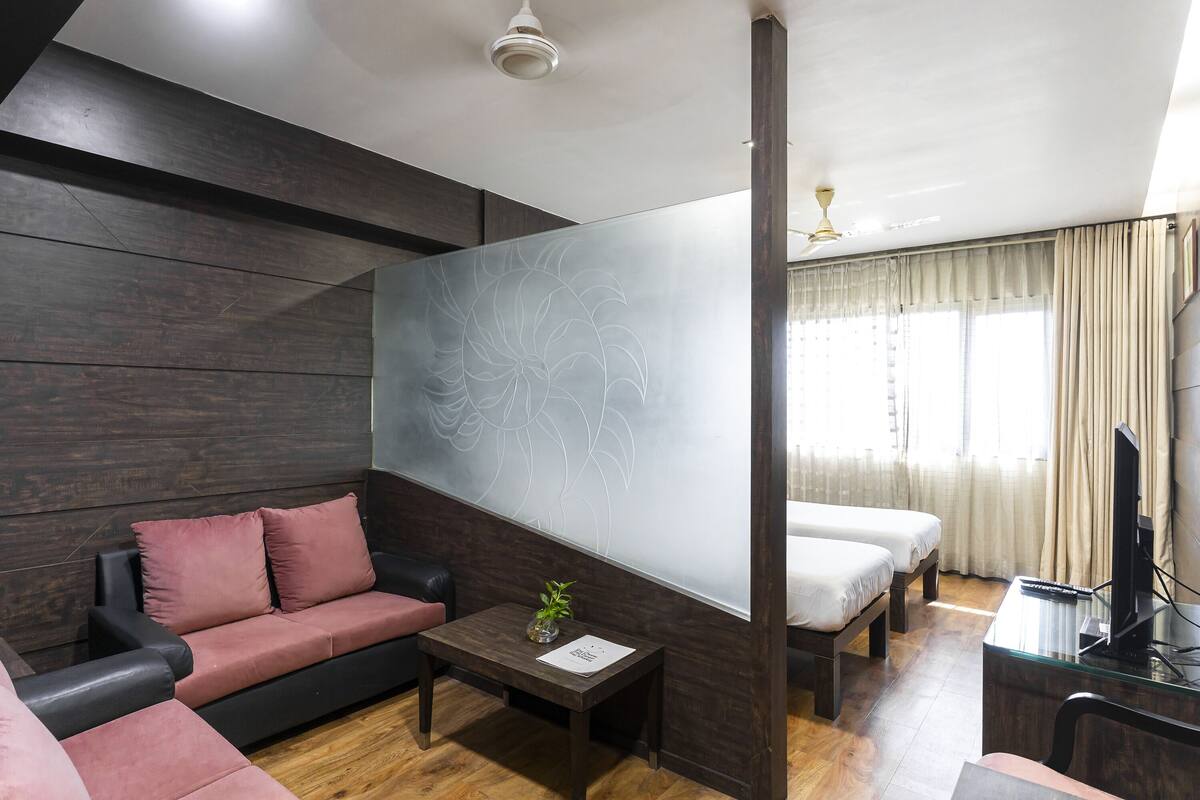 Una habitación de hotel moderna con una pared de paneles de madera oscura, un sofá, una cama, un escritorio con una computadora y un gran ventanal con cortinas.
