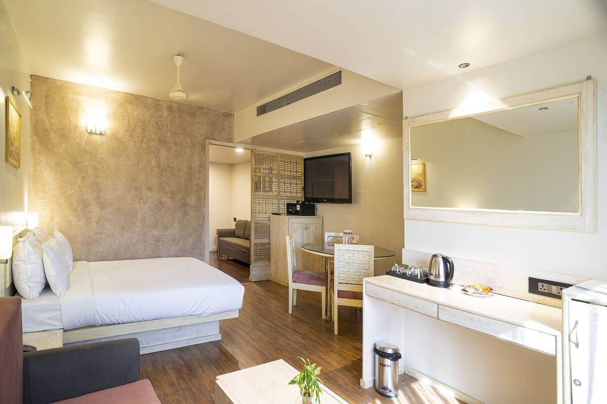 Habitación de hotel compacta con cama, un pequeño escritorio, una silla, kitchenette y televisión montada en la pared.