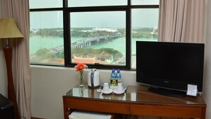 Habitación de hotel con un ventanal grande que ofrece vistas a un puente y al agua. Se ve un escritorio con un televisor, un florero con flores y algunas botellas.