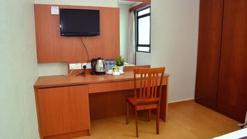A compact room with a wooden desk, a chair, a television mounted on the wall, a window with curtains, and a wardrobe.