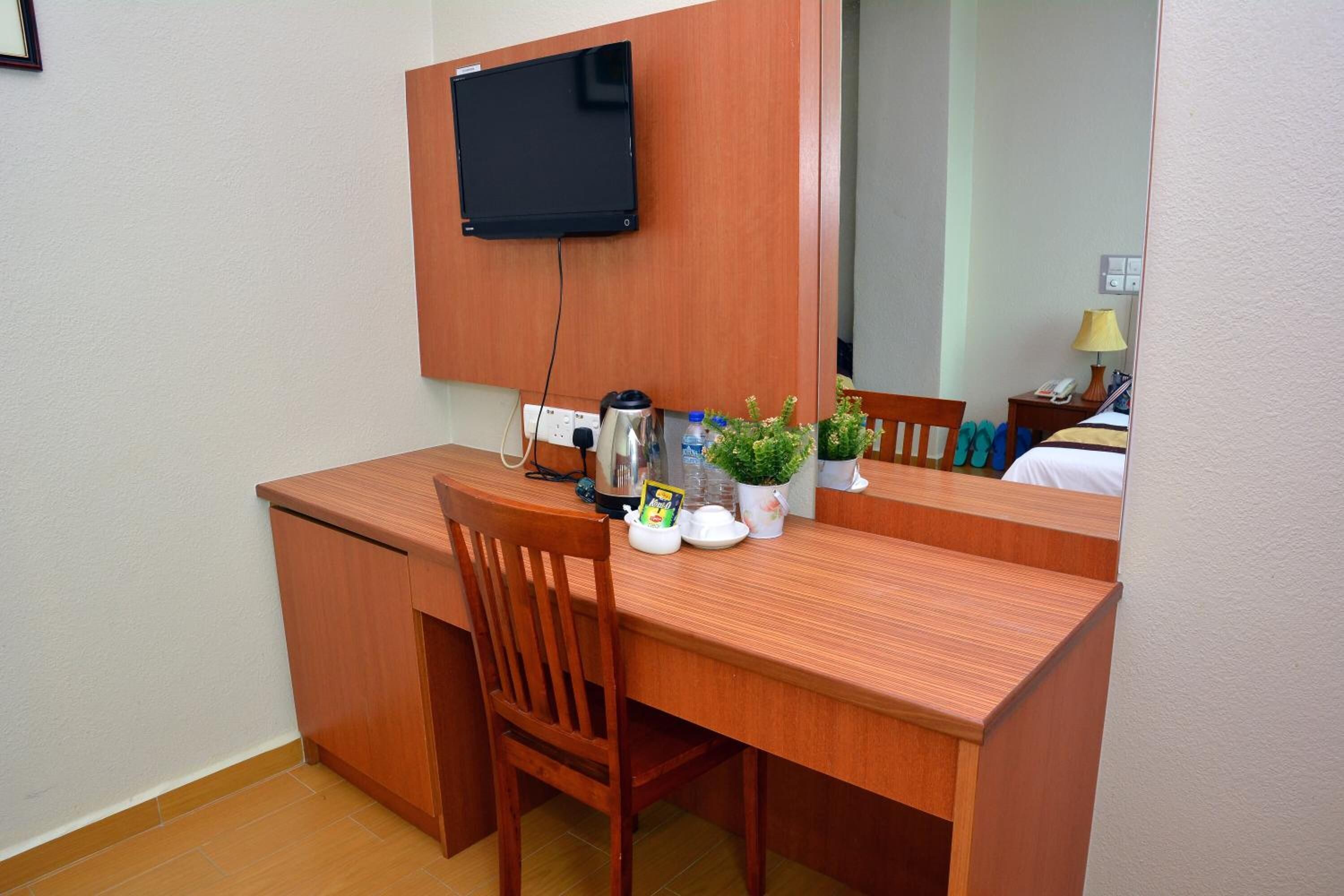 A compact hotel room with a wooden desk, a chair, a television mounted on the wall, a small potted plant, a kettle, and a bedside table with a lamp.