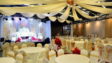 A banquet hall with round tables, chairs with golden sashes, and a stage with floral decorations.