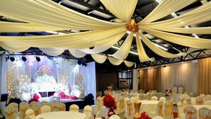 Un salón de banquetes con mesas redondas cubiertas con manteles blancos, sillas con fundas blancas y cintas doradas, y un escenario con decoración floral.