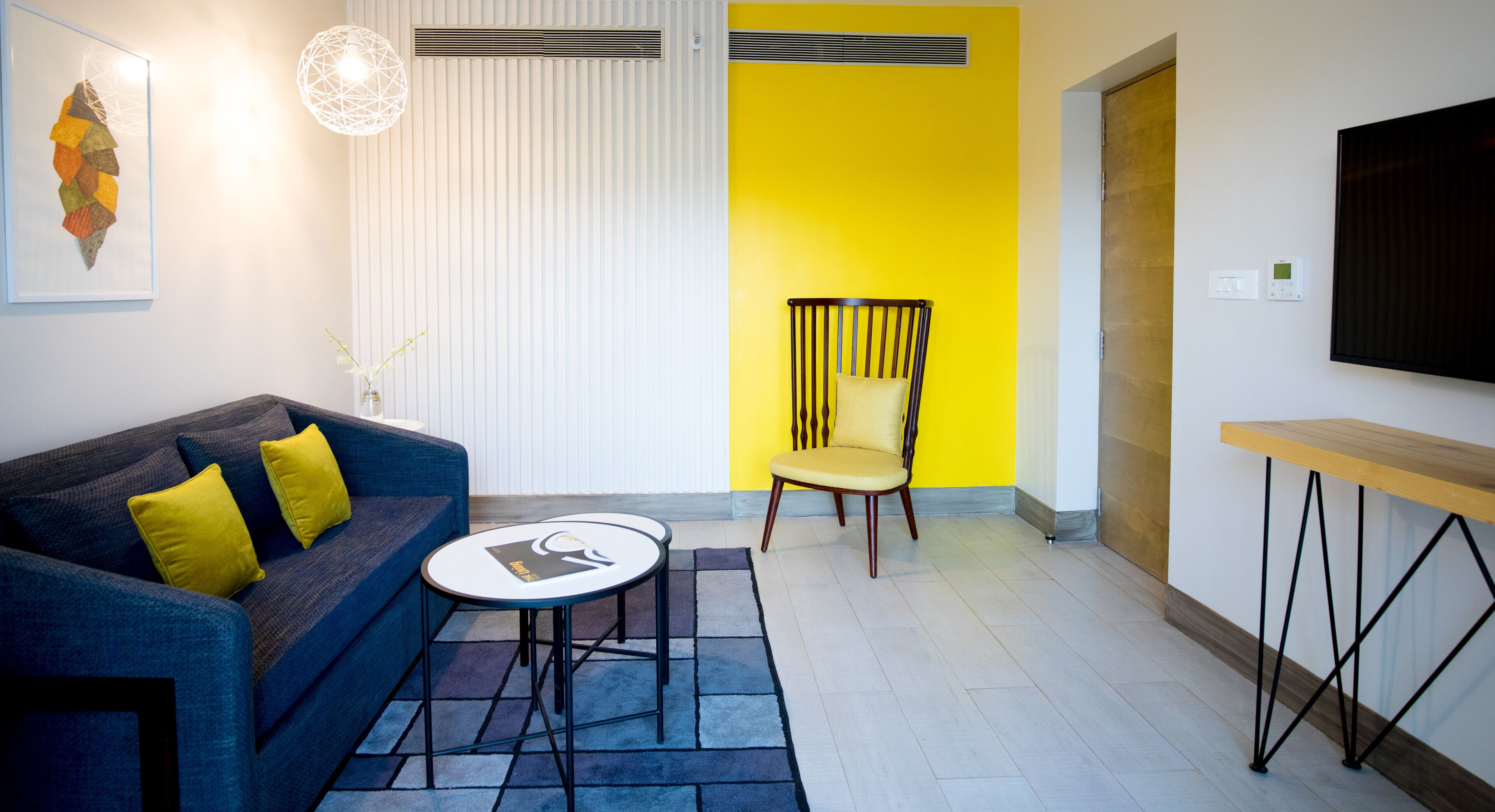 Una sala de estar moderna con un sofá azul, una pared de acento amarilla, una silla de madera, una mesita redonda y un televisor montado en la pared.