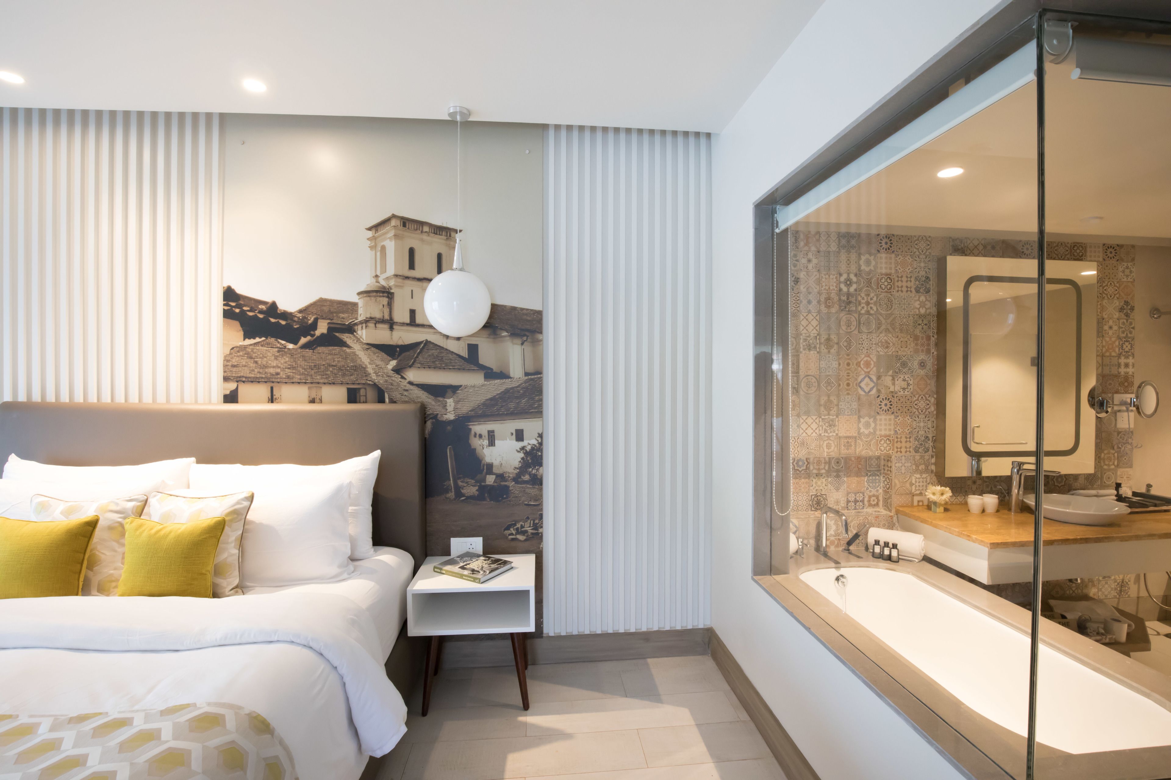 A modern hotel room with a large bed, a small bedside table, a large window with a view of buildings, and a bathroom with a bathtub and sink.