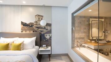 A modern hotel room with a large bed, a small bedside table, a large window with a view of buildings, and a bathroom with a bathtub and sink.