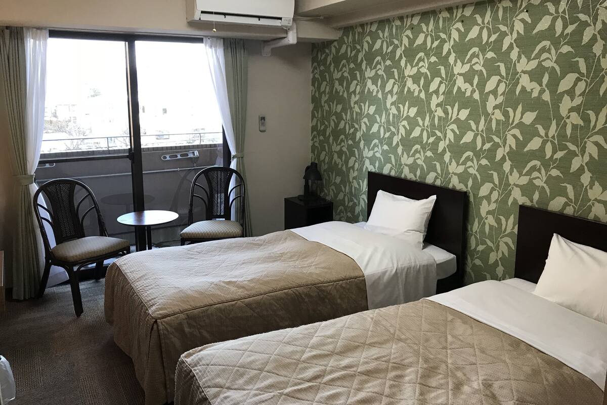 A hotel room with two beds, a small table, two chairs, an air conditioner, and a window with curtains.