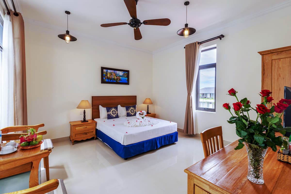 A hotel room with a bed, a dining table, chairs, a vase with flowers, a ceiling fan, and a painting on the wall.