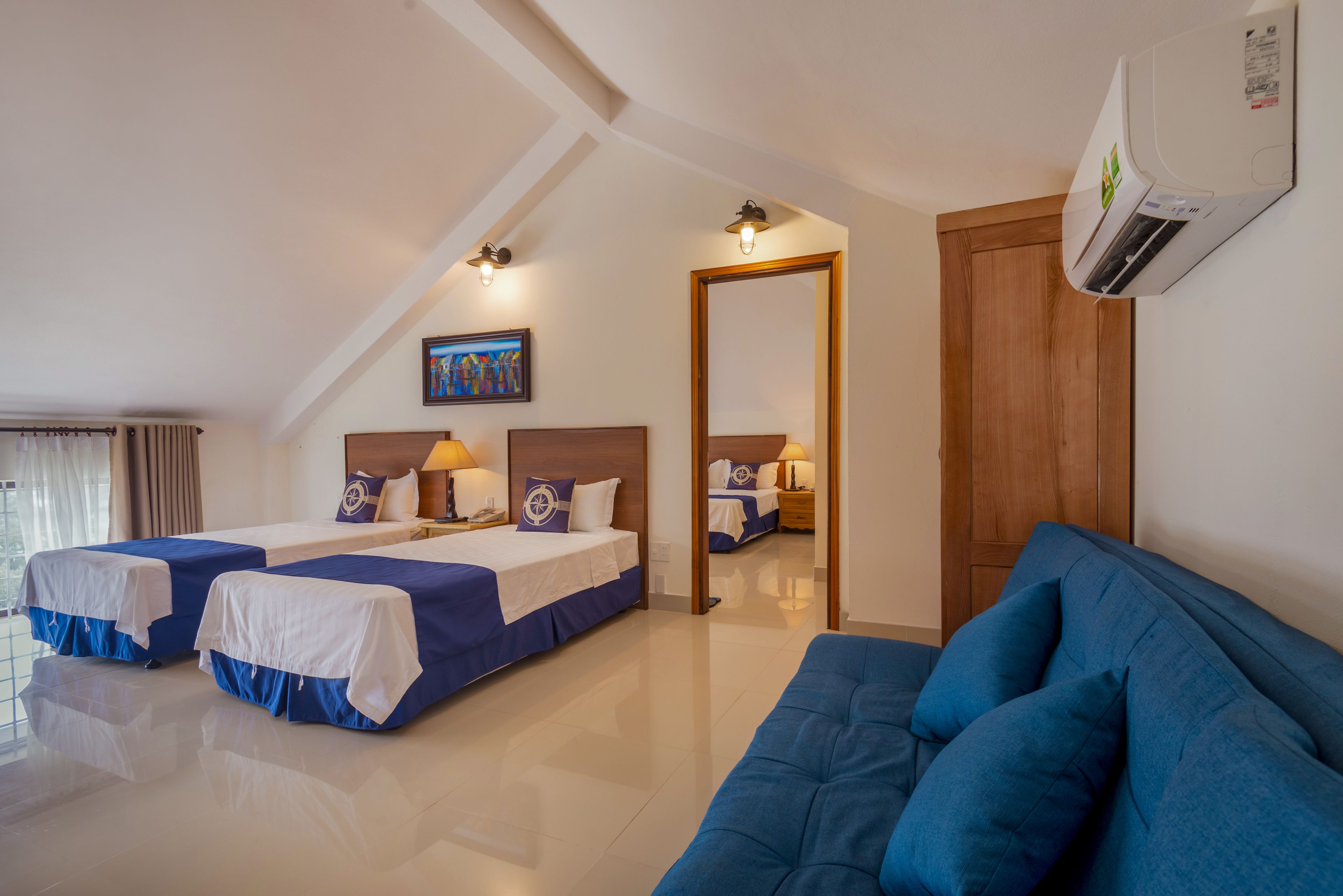 A hotel room with two beds, a blue sofa, a painting on the wall, and an air conditioning unit.