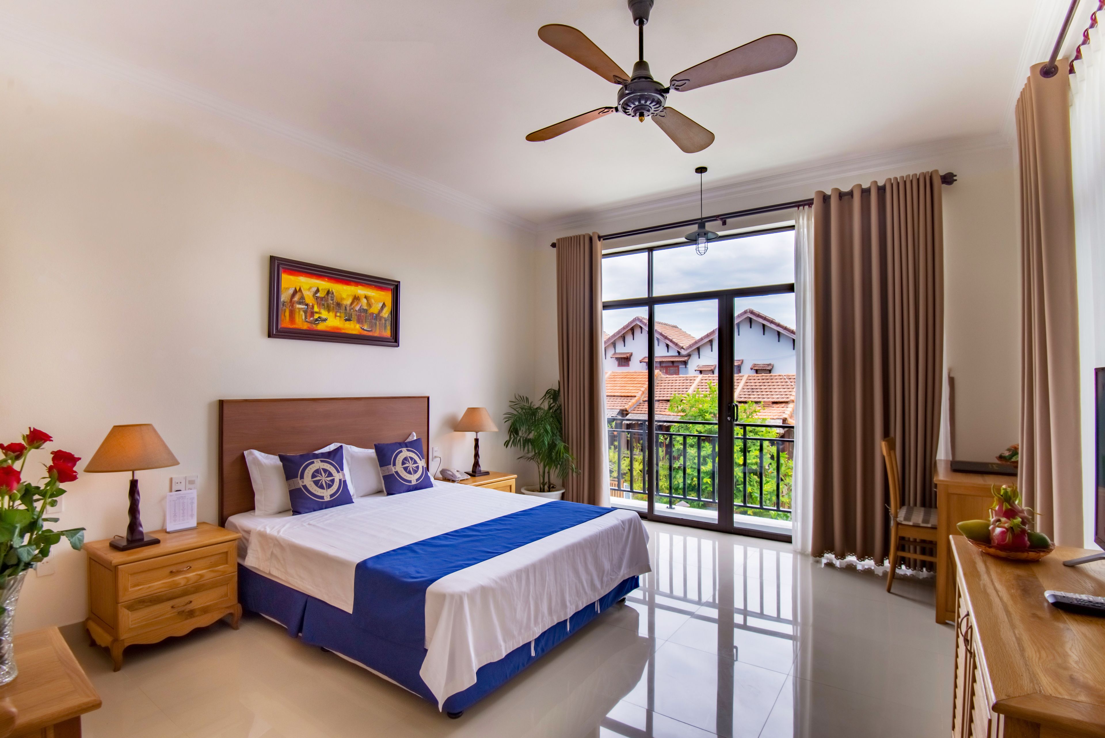 A hotel room with a bed, nightstands, a ceiling fan, a window with curtains, and a balcony with a view of buildings.