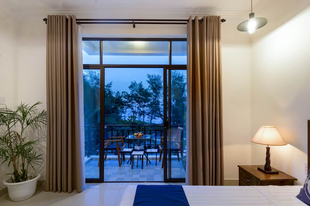 A hotel room with a bed, a nightstand with a lamp, a potted plant, and a view of an outdoor dining area through a sliding glass door.