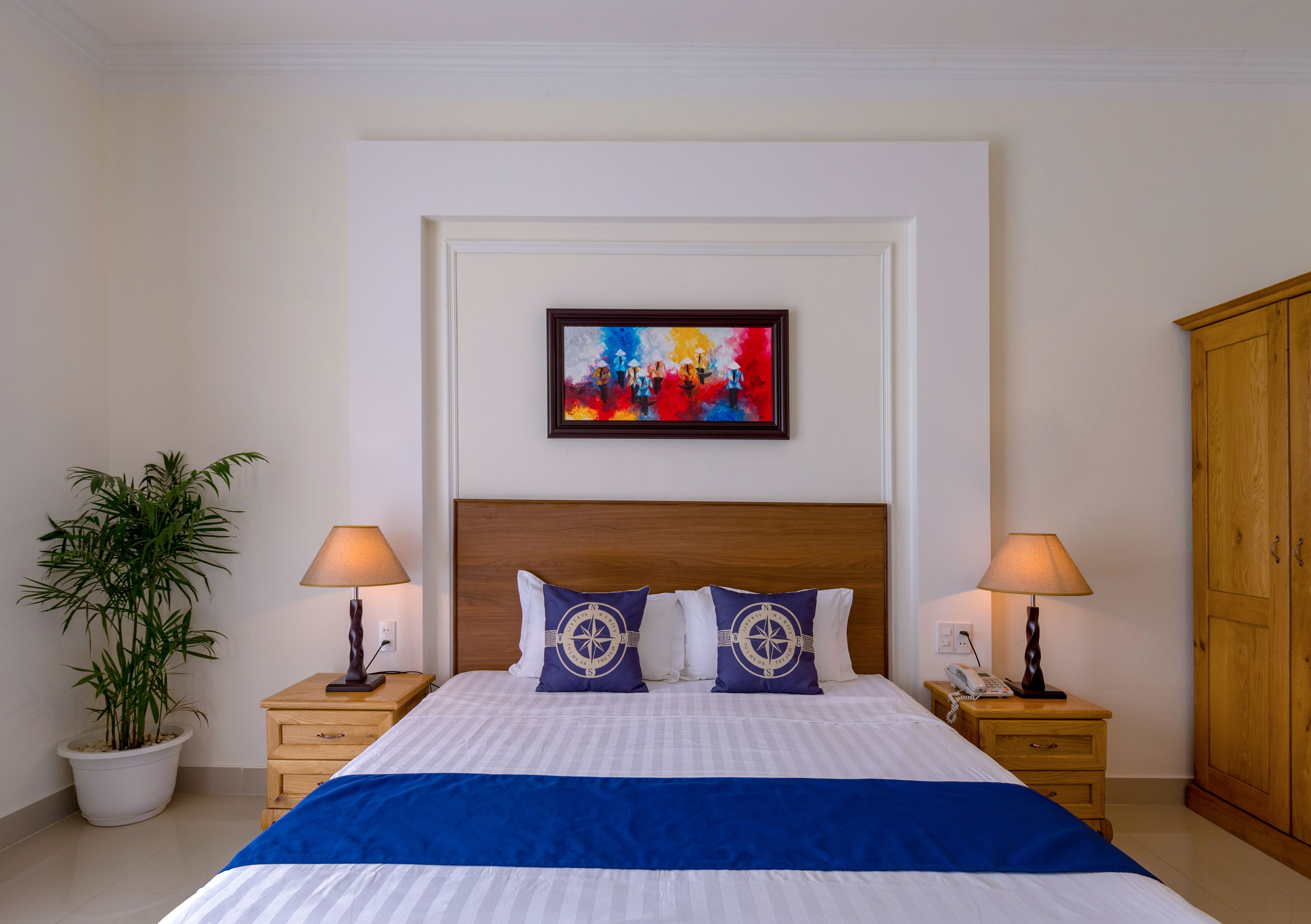 A neatly arranged hotel room with a large bed, two bedside tables, a potted plant, a wooden wardrobe, and a colorful abstract painting on the wall.