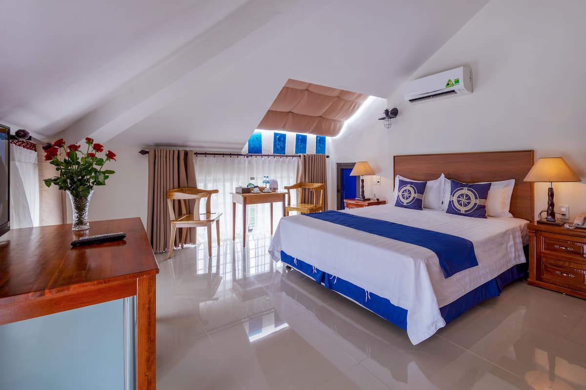 A hotel room with a bed, a desk, a chair, a table, a vase with flowers, and an air conditioning unit.