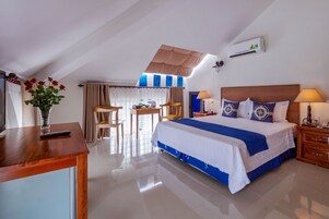 Una habitación de hotel con cama, escritorio, silla, mesa, un jarrón con flores y un equipo de aire acondicionado.