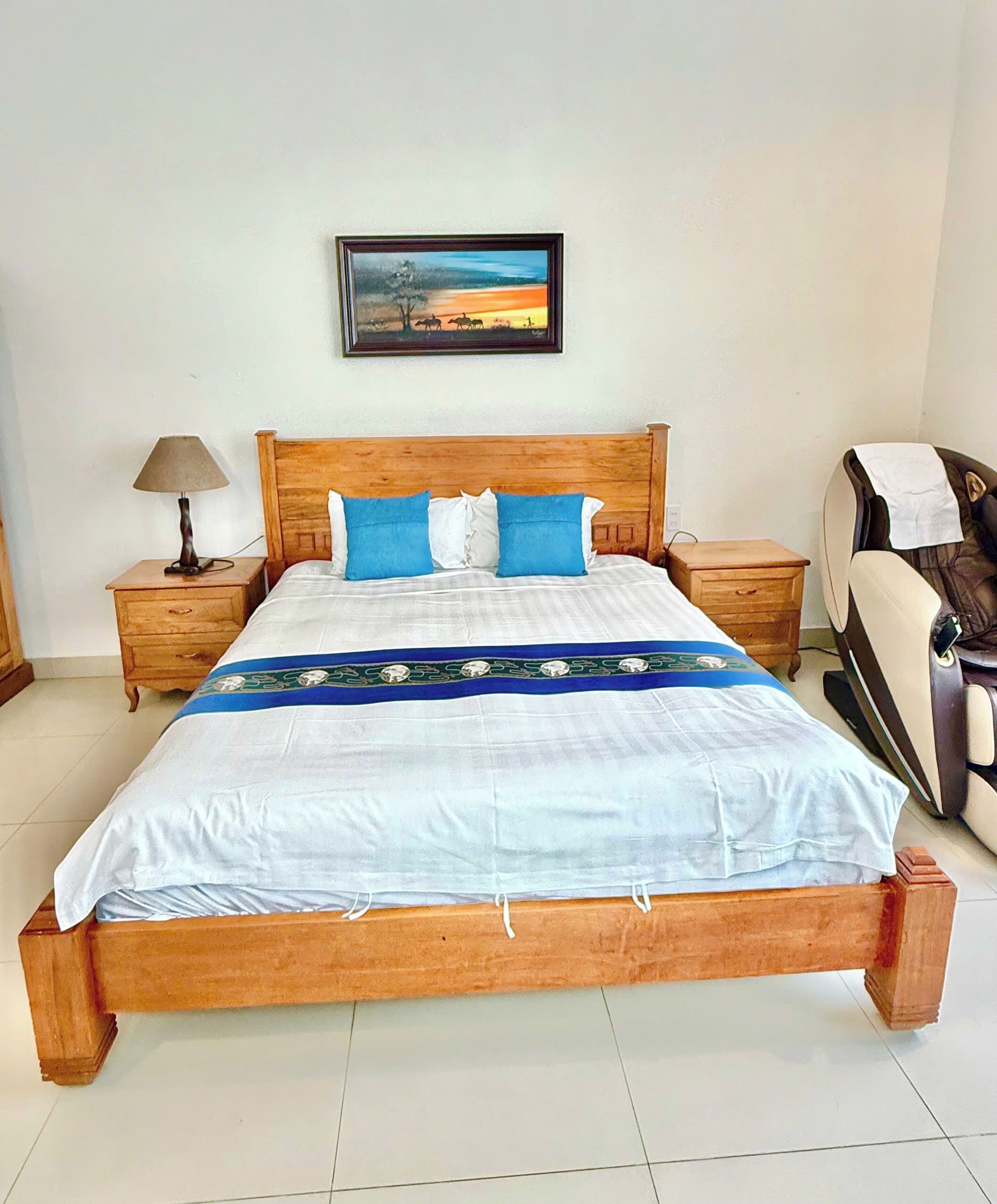 Una cama de madera con colcha blanca y azul, dos mesitas de noche con lámparas, una silla de masaje y un cuadro en la pared.