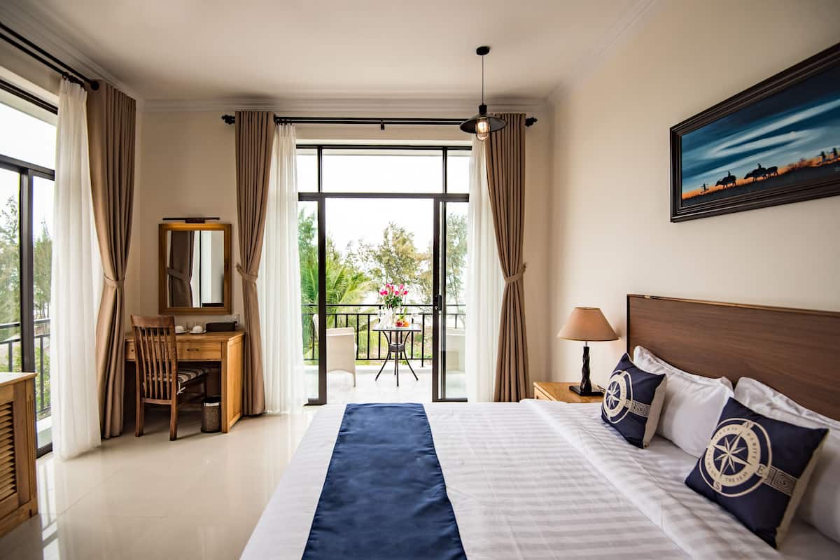 A hotel room with a bed, a desk, a chair, a mirror, a nightstand with a lamp, and a balcony with a view of trees and a building.