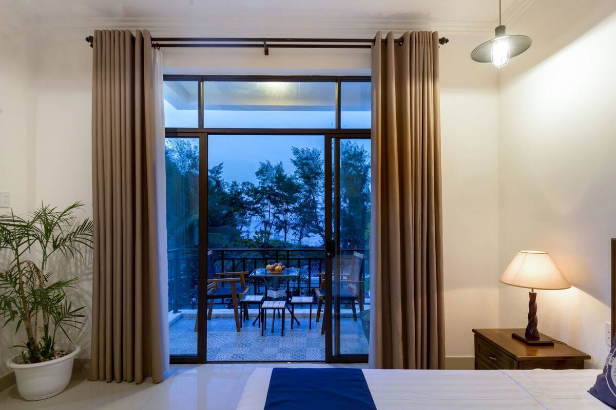 A hotel room with a bed, a nightstand with a lamp, a potted plant, and a view of an outdoor seating area with tables and chairs.