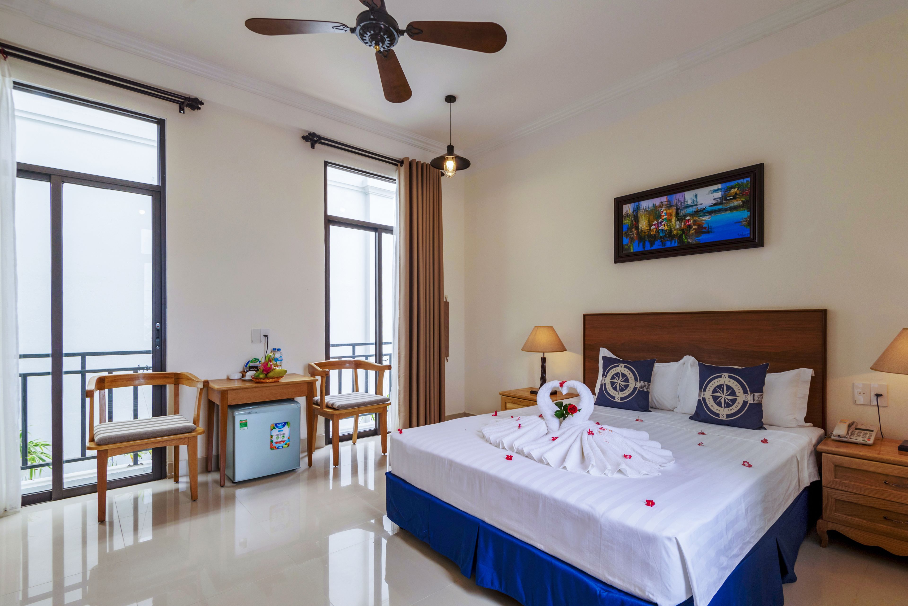 A hotel room with a bed, a desk, a chair, a mini-fridge, a ceiling fan, a painting, and a nightstand with a lamp.