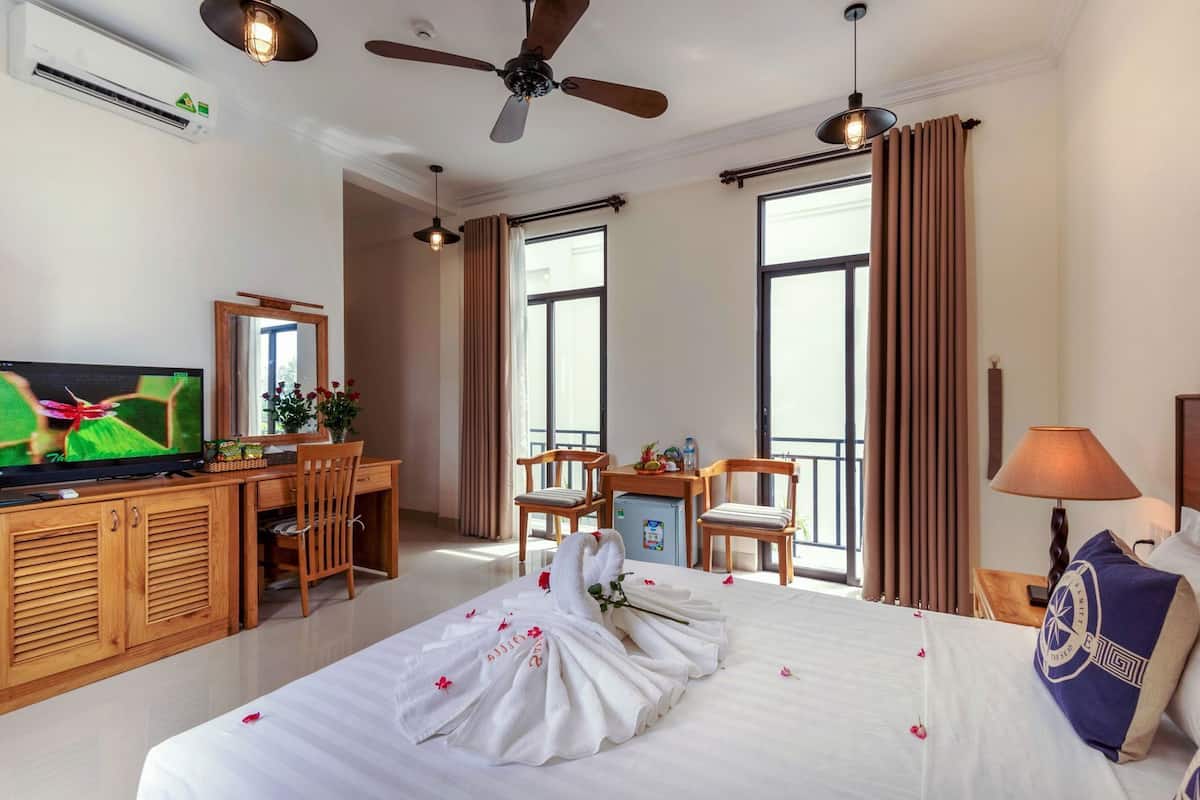 A hotel room with a bed, a desk with a television, a balcony, and large windows.