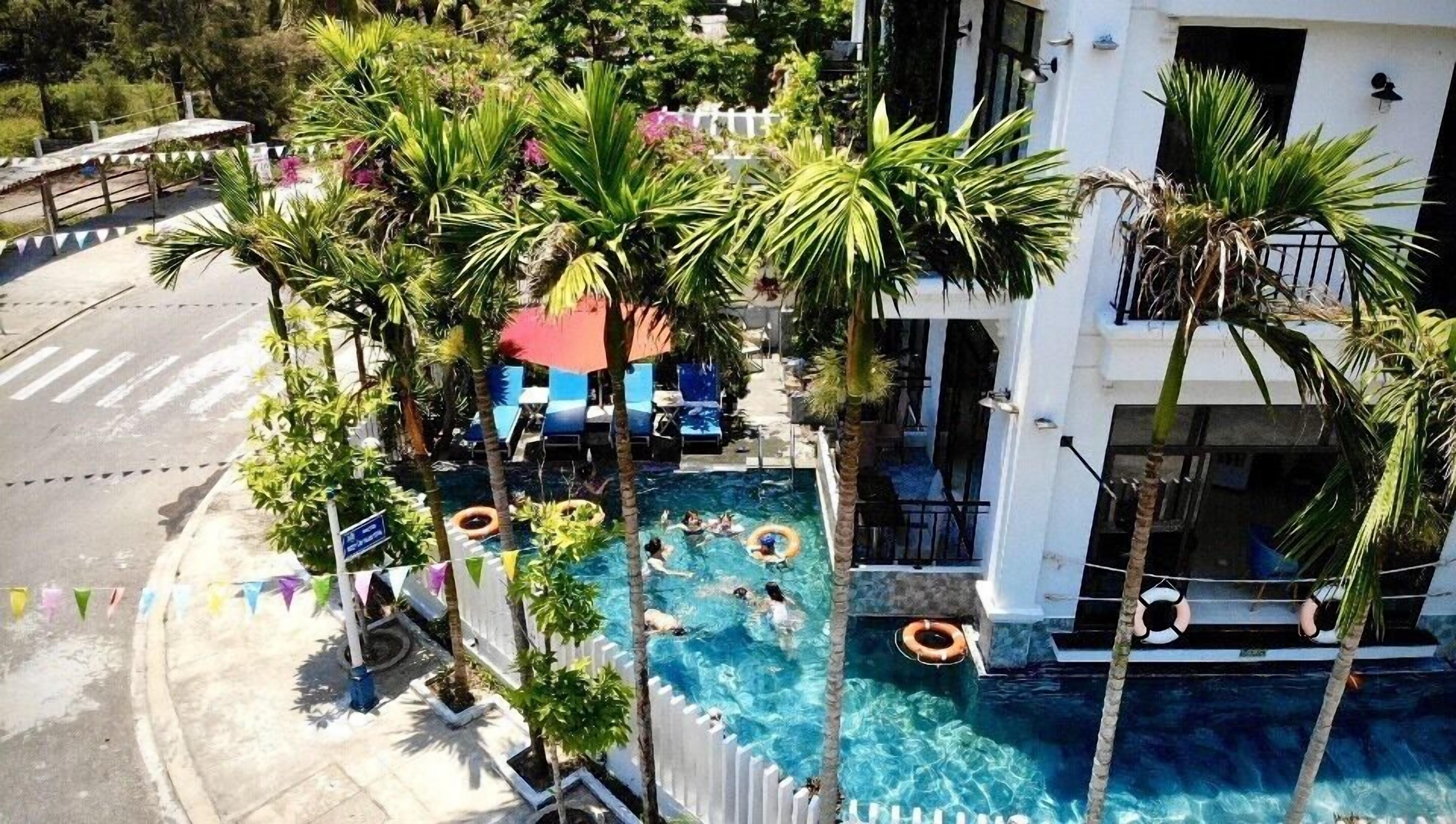 An aerial view of a hotel with a swimming pool surrounded by palm trees, lounge chairs, and a white fence.