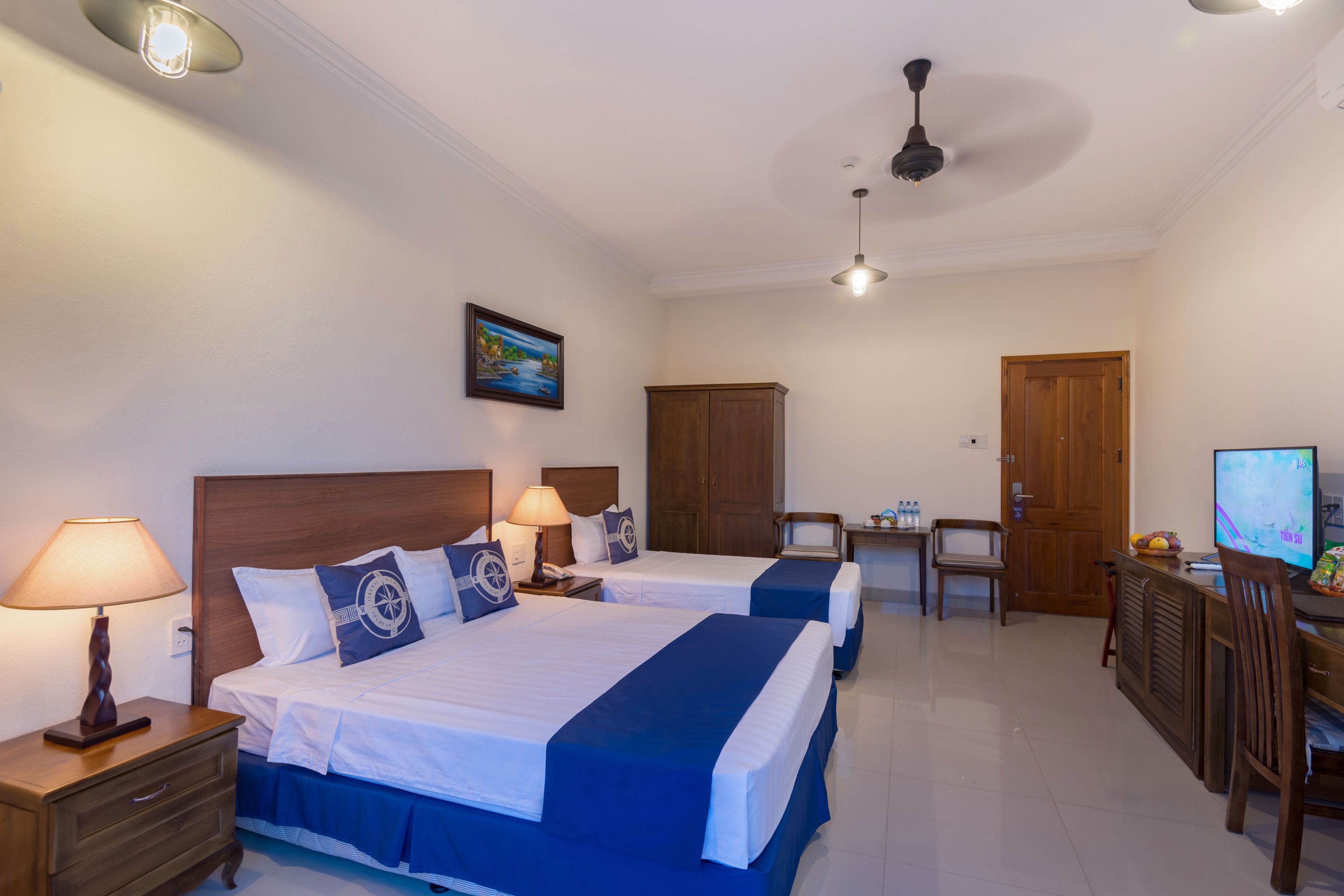 A hotel room with two beds, a desk, a television, and a ceiling fan.