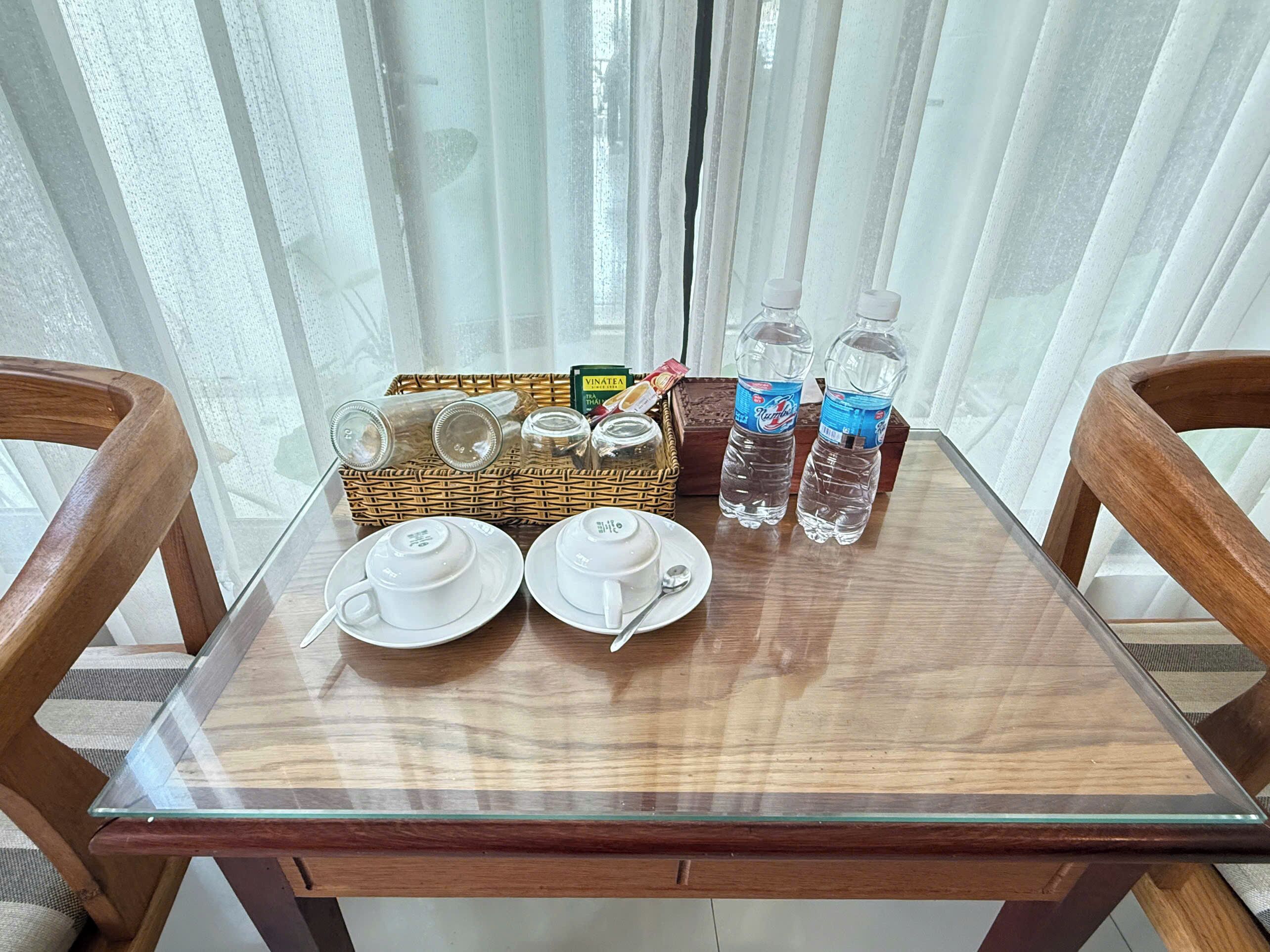 A wooden table with two white teacups and saucers, a wicker basket with jars and a box, and two water bottles.