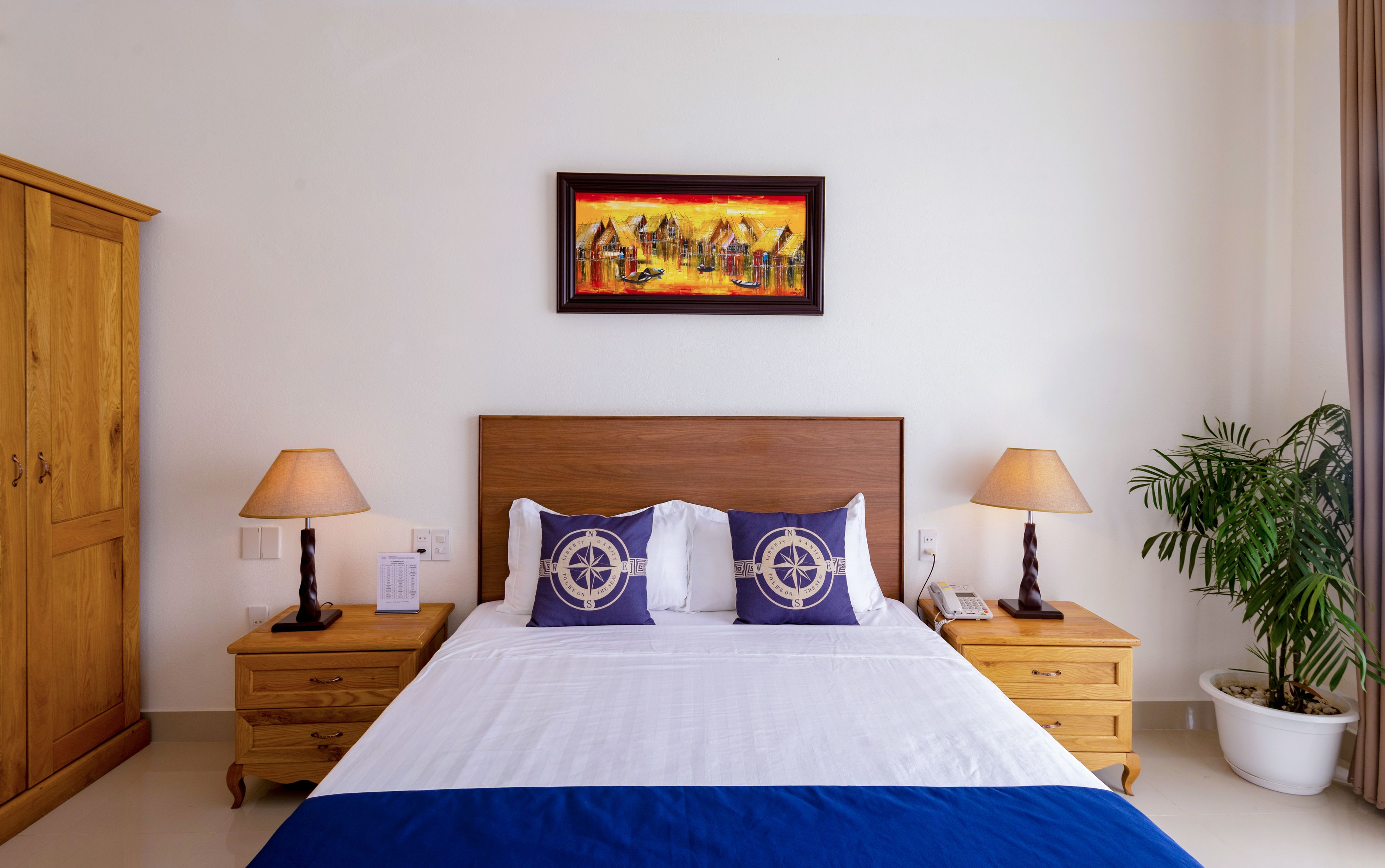 A hotel room with a large bed, two bedside tables with lamps, a wooden wardrobe, a painting on the wall, and a potted plant.