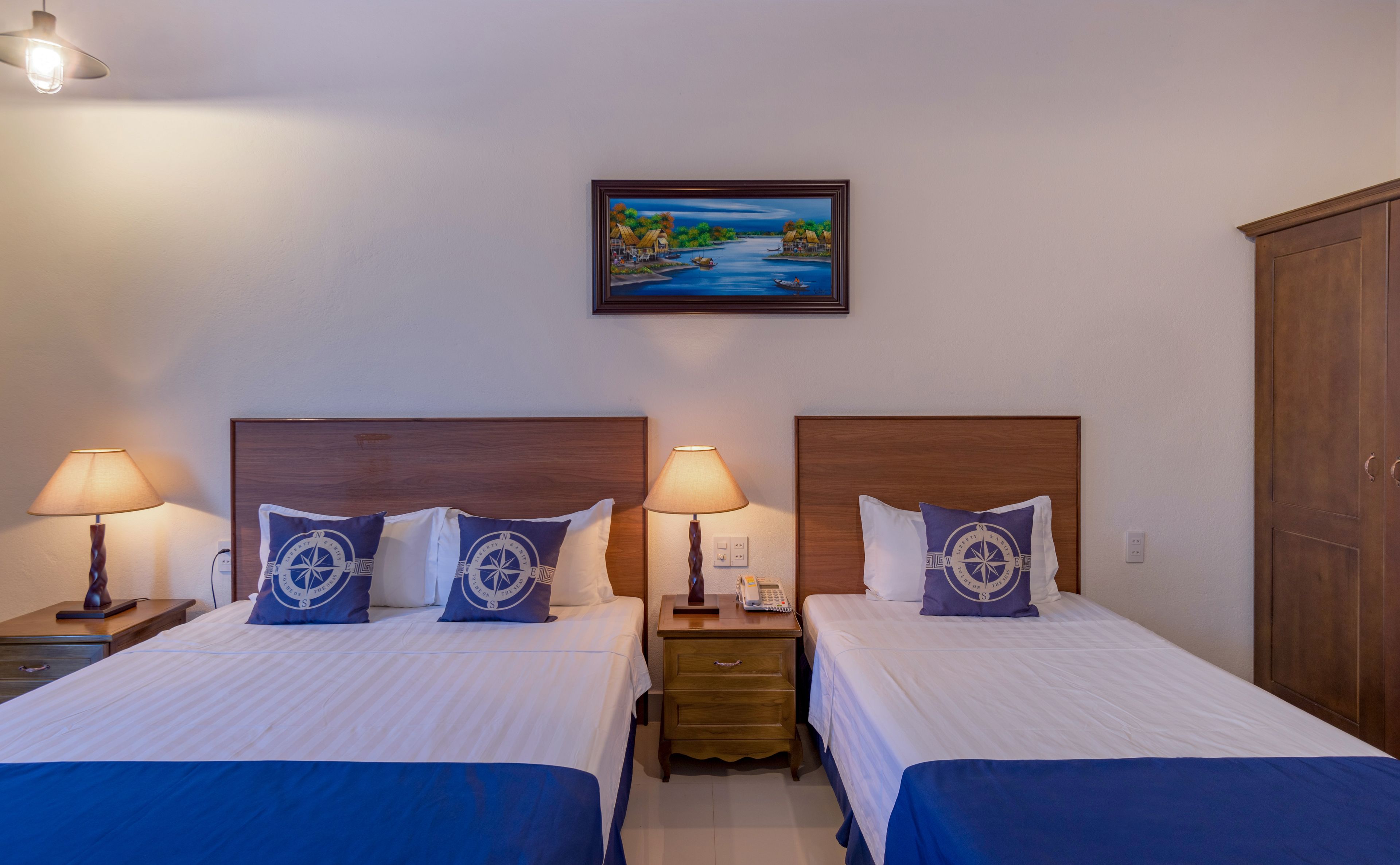 A hotel room with two beds, each with a wooden headboard, a nightstand with a lamp, and a painting of a river scene on the wall.