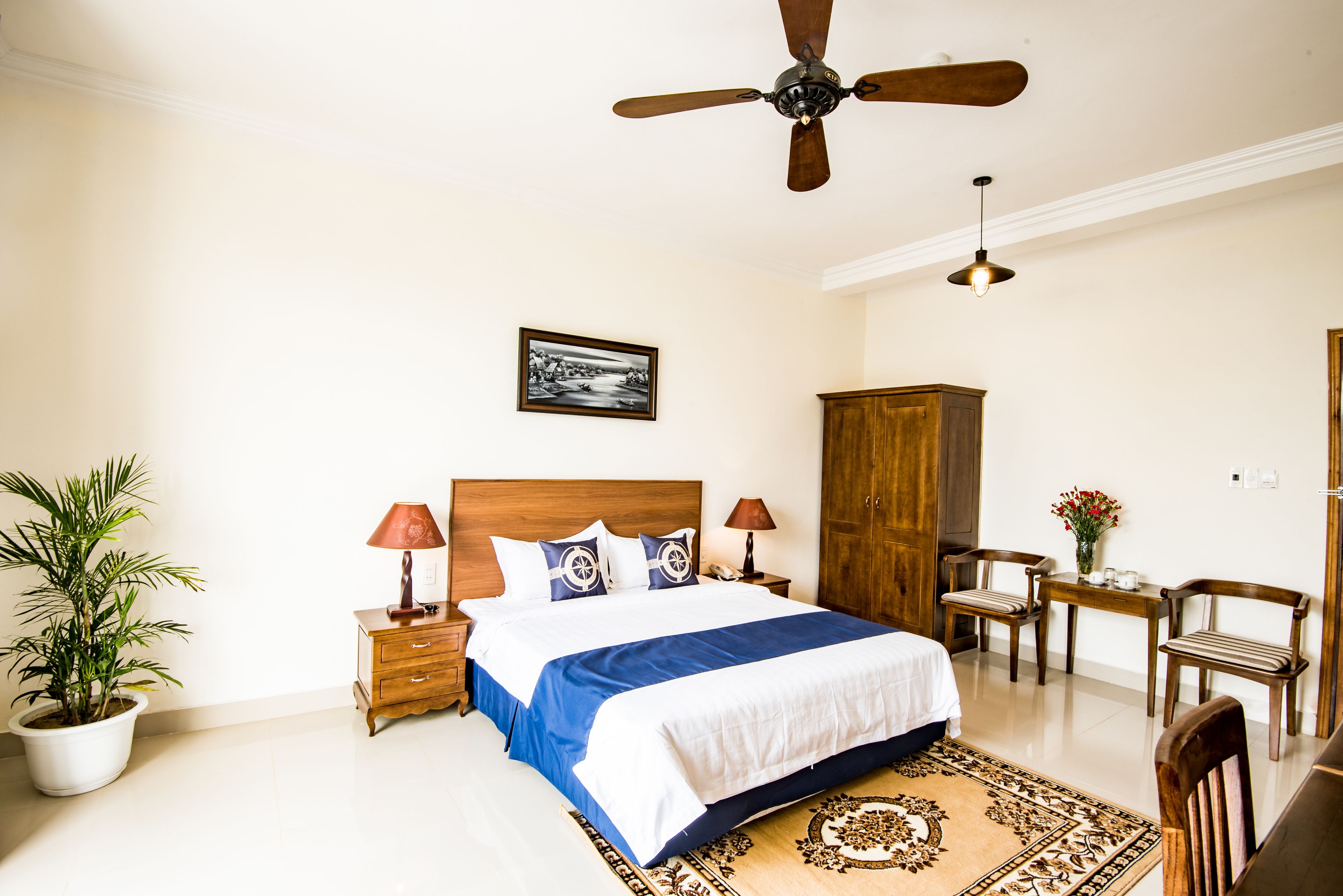 A bedroom with a bed, nightstands, a wooden wardrobe, a ceiling fan, a desk with a chair, and a potted plant.