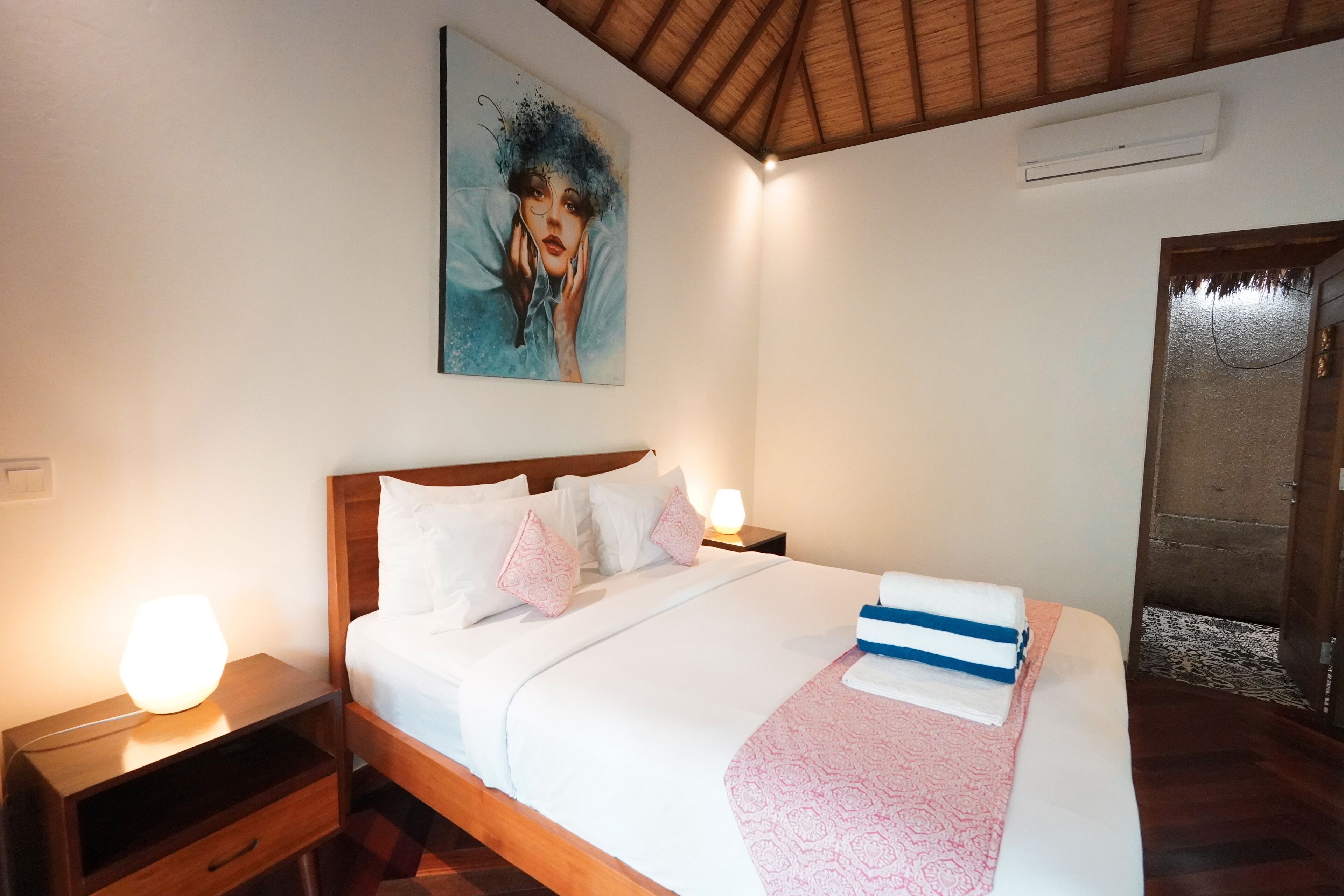 A neatly made bed with white linens and pink patterned pillows, a bedside lamp, and a painting on the wall.