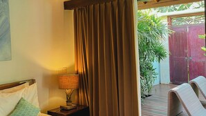 Un cuarto de hotel con cama, mesita de noche con lámpara, ventana con cortinas y vista a una terraza al aire libre.