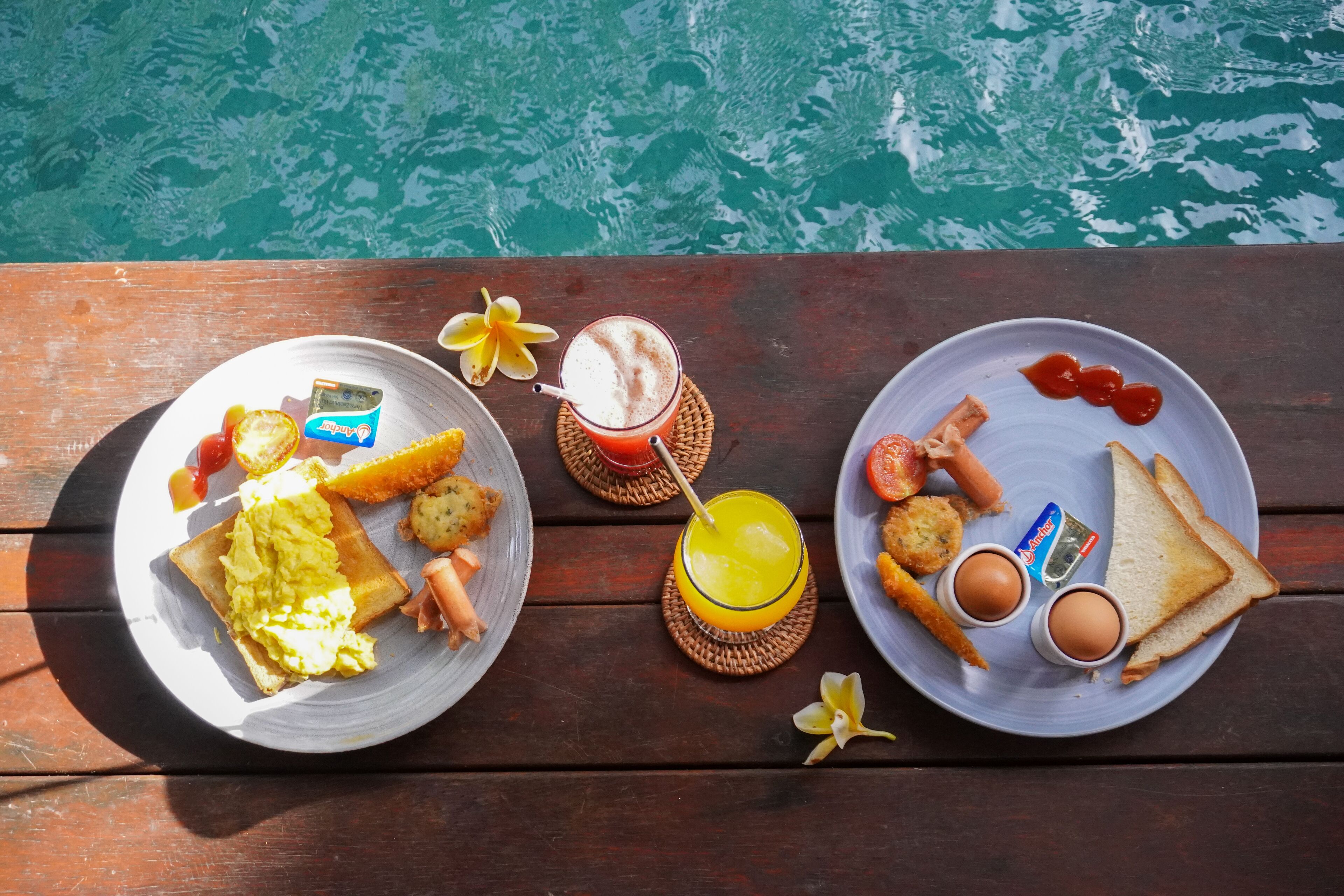 Une table en bois, dressée avec des aliments pour le petit-déjeuner, notamment des toasts, des œufs et une boisson, près d’une étendue d’eau.