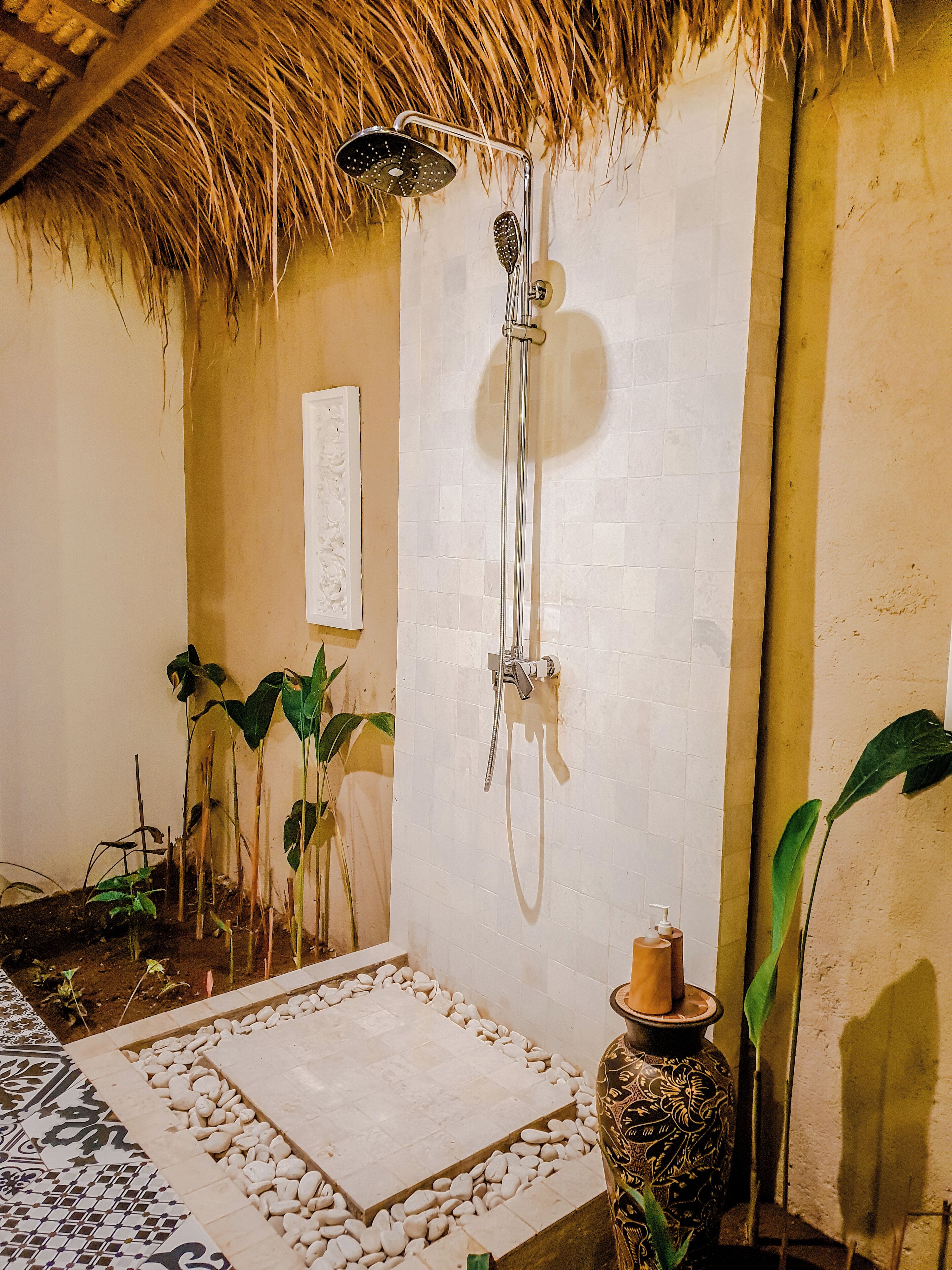 A bathroom with a shower, a tiled floor, a thatched roof, and potted plants.